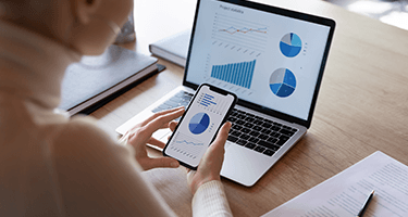 A person sits at a desk holding their phone showing a graph, while their laptop sits on the desk showing more charts and graphs.